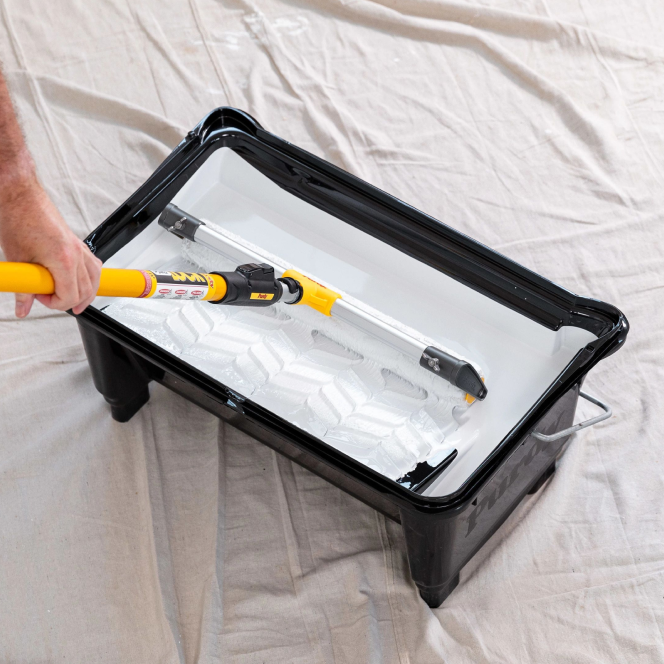 Painter rolling out white paint with a large roller in an eighteen-inch paint tray.